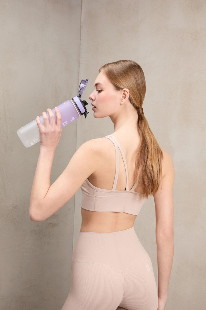 A woman in a beige sports bra and matching leggings drinks from a translucent purple water bottle with a black and purple flip-top lid, set against a textured gray concrete wall.