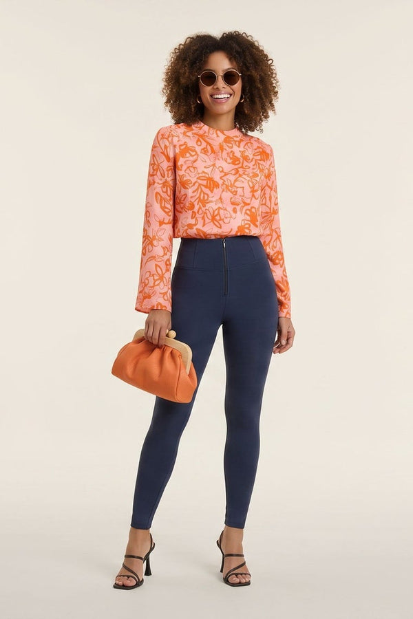 Woman wearing an orange patterned top and blue pants, holding an orange handbag, on a white background.