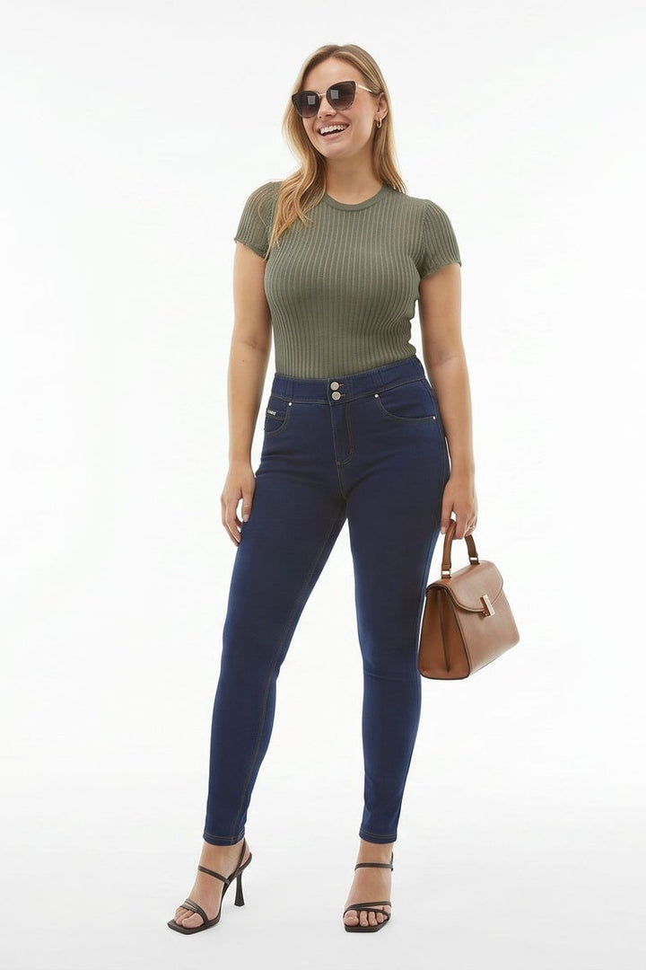Woman wearing blue jeans and a green top, holding a brown handbag, on a white background.