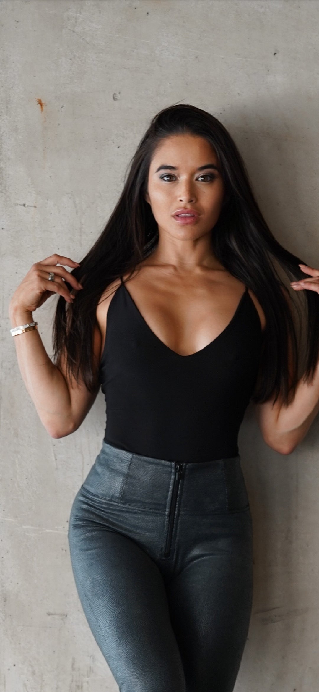 Woman wearing a black tank top and blue jeans against a gray concrete wall.