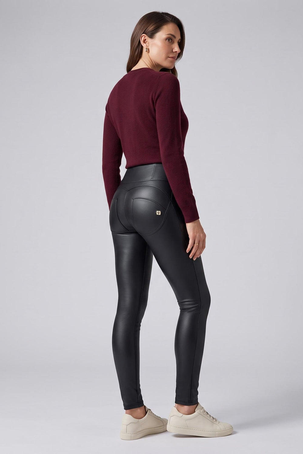 A woman stands in profile wearing black high-waisted faux-leather leggings, a dark burgundy long-sleeve top, and off-white sneakers against a light gray studio background.