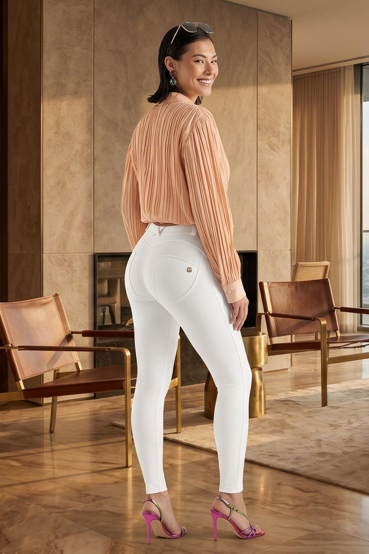 Woman in a peach blouse and white pants standing in a modern living room with large windows.