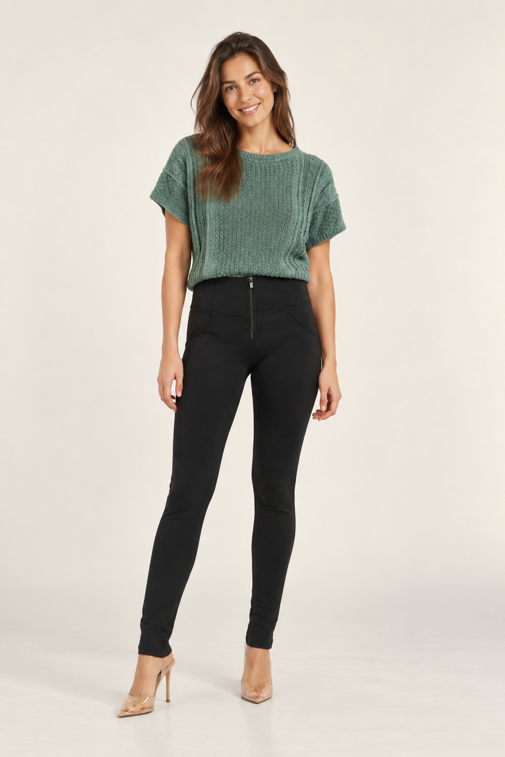 A woman modeling a short-sleeve textured sage green top and black skinny pants with a front zipper, paired with nude high heels, against a plain light beige studio background.