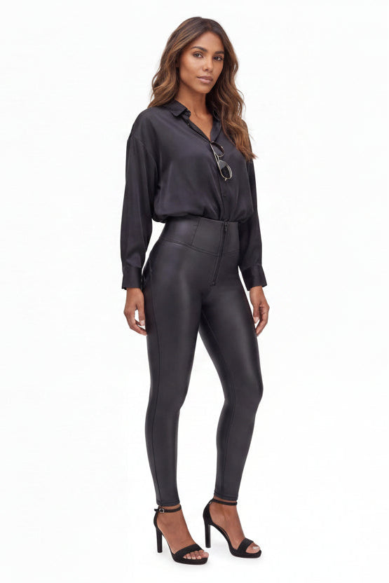 A woman modeling a black long-sleeve collared shirt paired with high-waisted, shiny black faux-leather pants and black heeled sandals, photographed against a plain white studio background.
