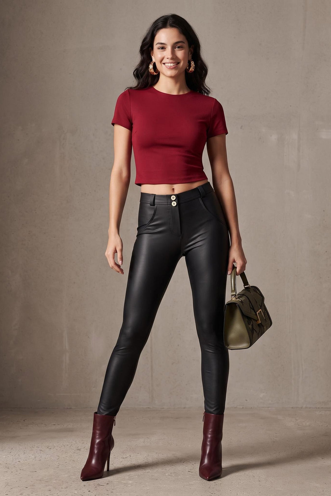 A woman stands against a textured beige studio wall wearing a fitted short-sleeve crop top in deep burgundy, high-waisted black faux-leather pants, burgundy ankle boots, and holding a small taupe handbag.