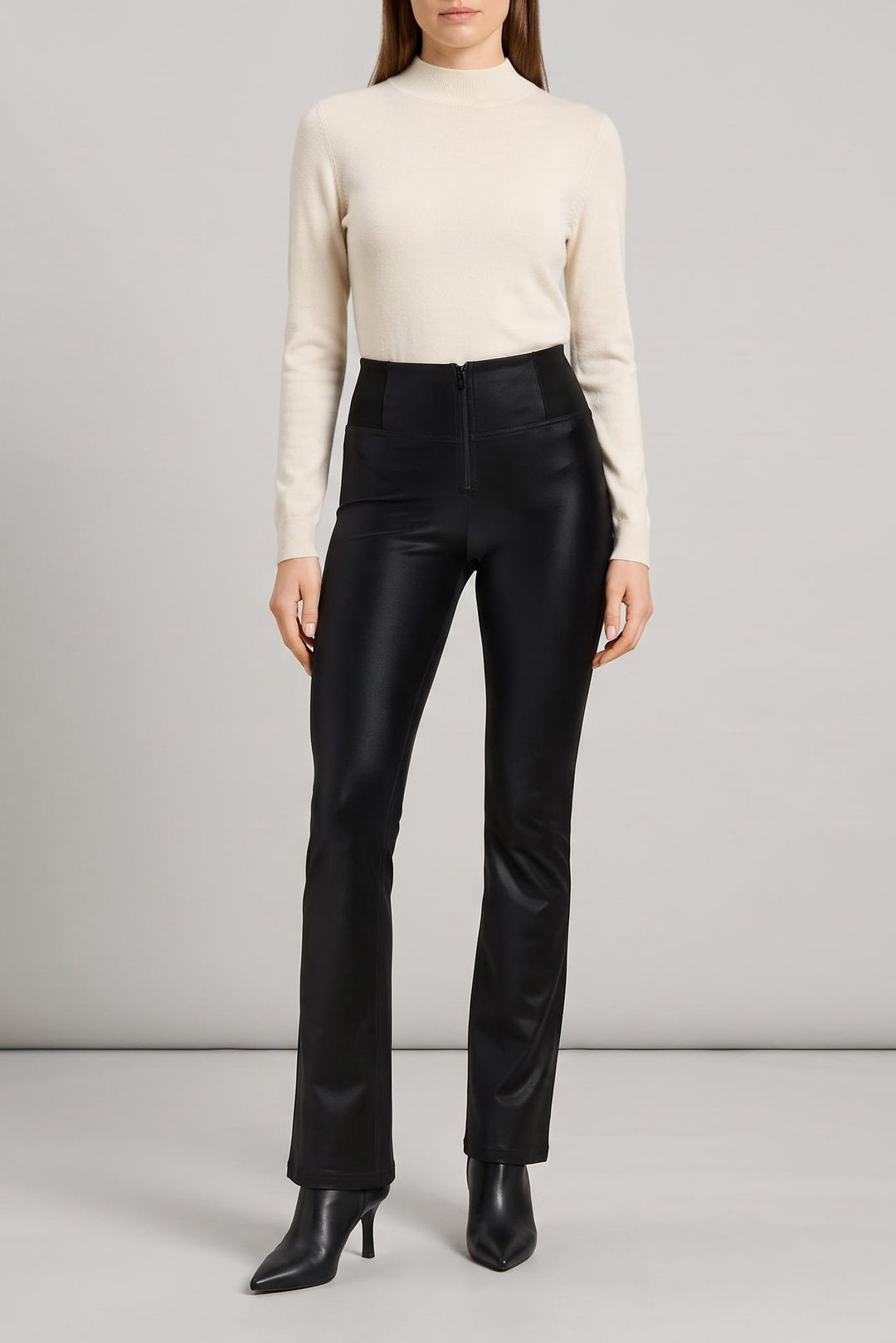 A model wearing a cream mock-neck knit sweater and high-waisted black leather flare pants with black pointed-toe heeled boots, standing against a light gray studio background.