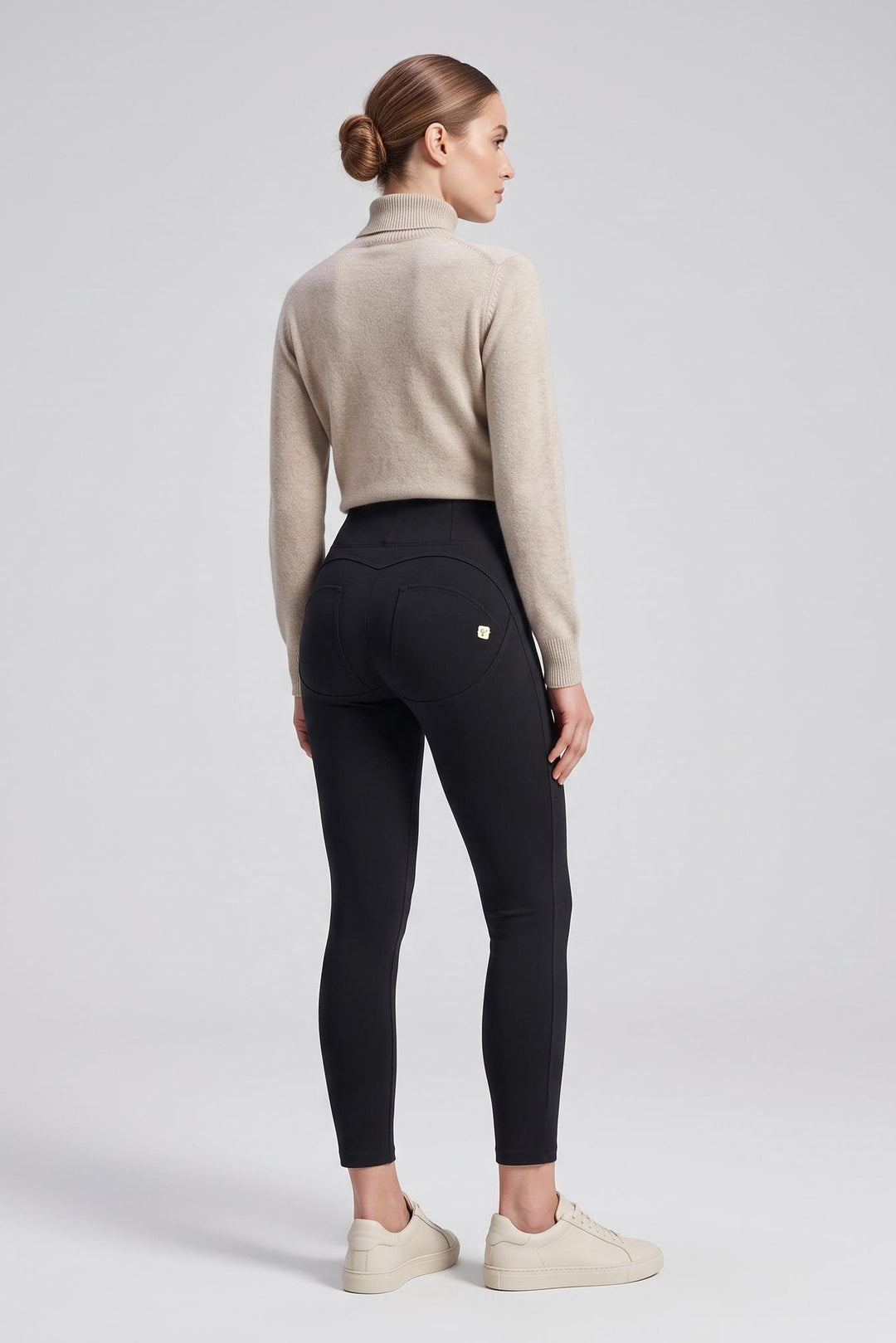 A woman stands in profile wearing black high-waisted leggings, a light beige cropped turtleneck sweater, and off-white sneakers against a light gray studio background.