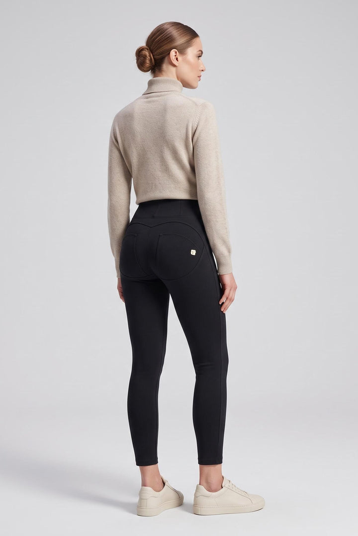 A woman stands in profile wearing black high-waisted leggings, a light beige cropped turtleneck sweater, and off-white sneakers against a light gray studio background.