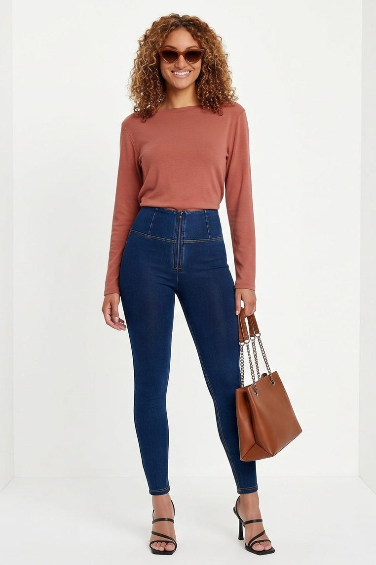 Woman wearing blue jeans and a brown top, holding a brown bag on a white background