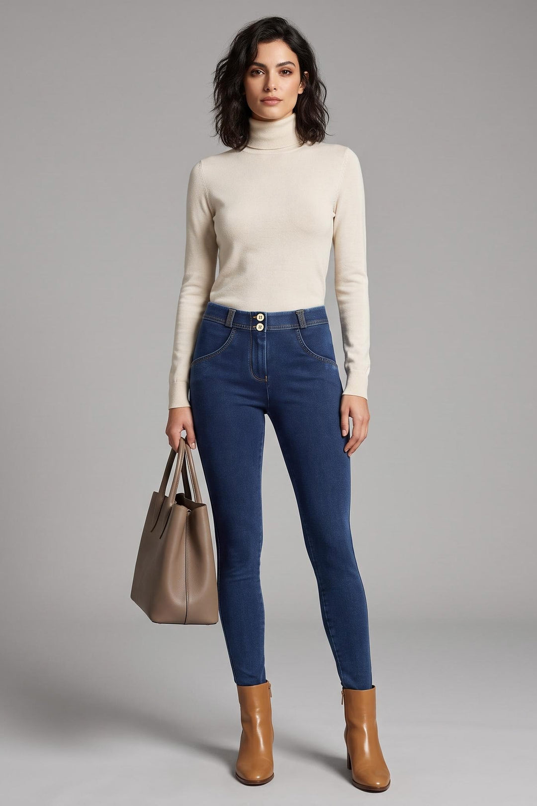 A woman modeling a cream ribbed turtleneck sweater, blue skinny jeans, and brown ankle boots, holding a taupe tote bag against a neutral gray studio background.