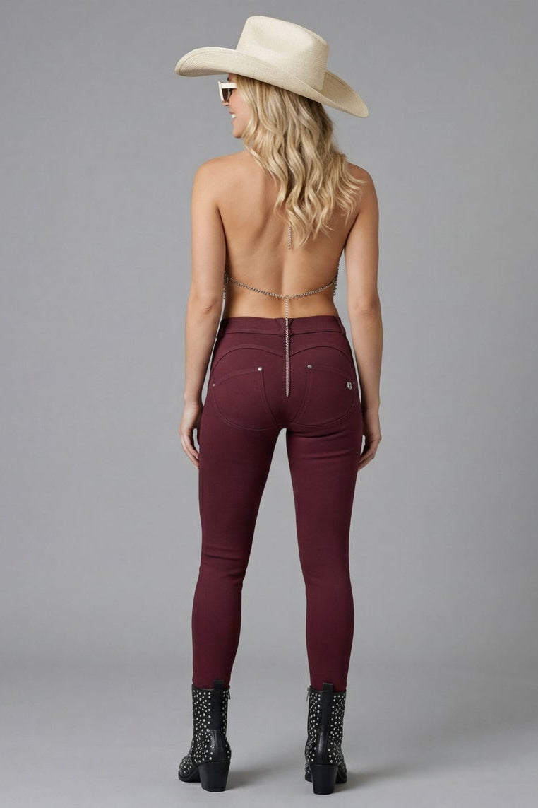 Woman wearing burgundy pants, fishnet top, and cowboy hat on a gray background
