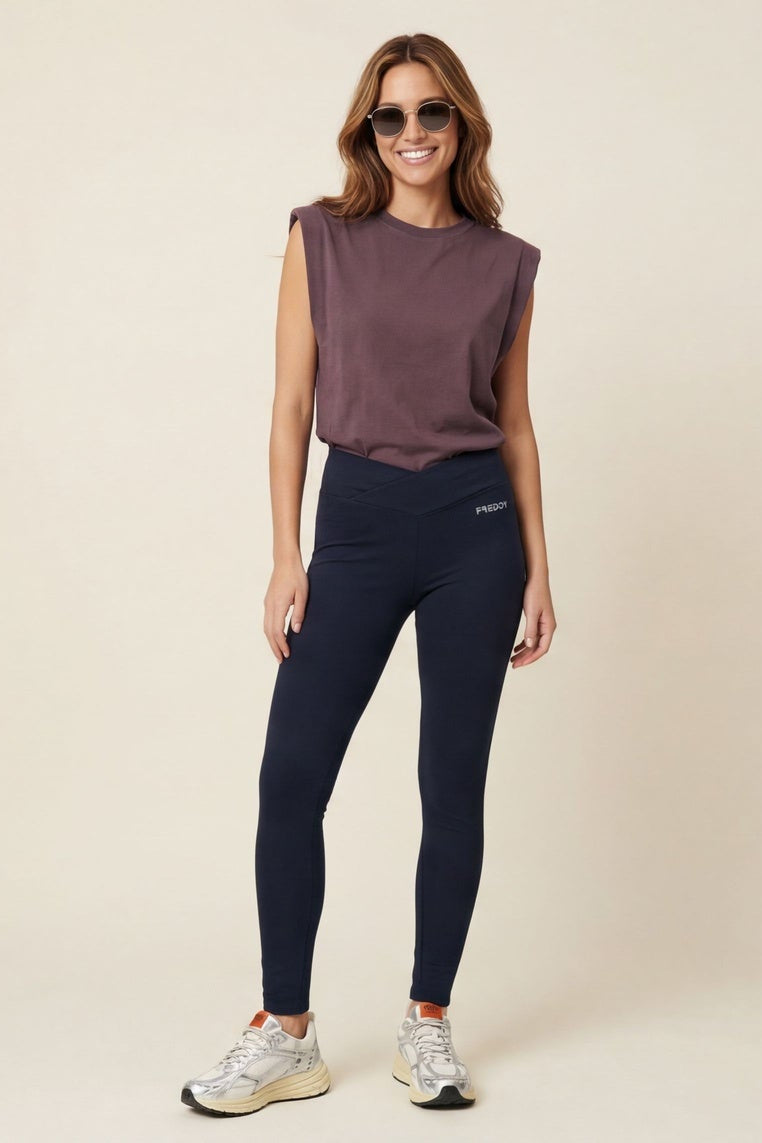 Woman wearing a purple sleeveless top and dark blue leggings standing against a beige background.