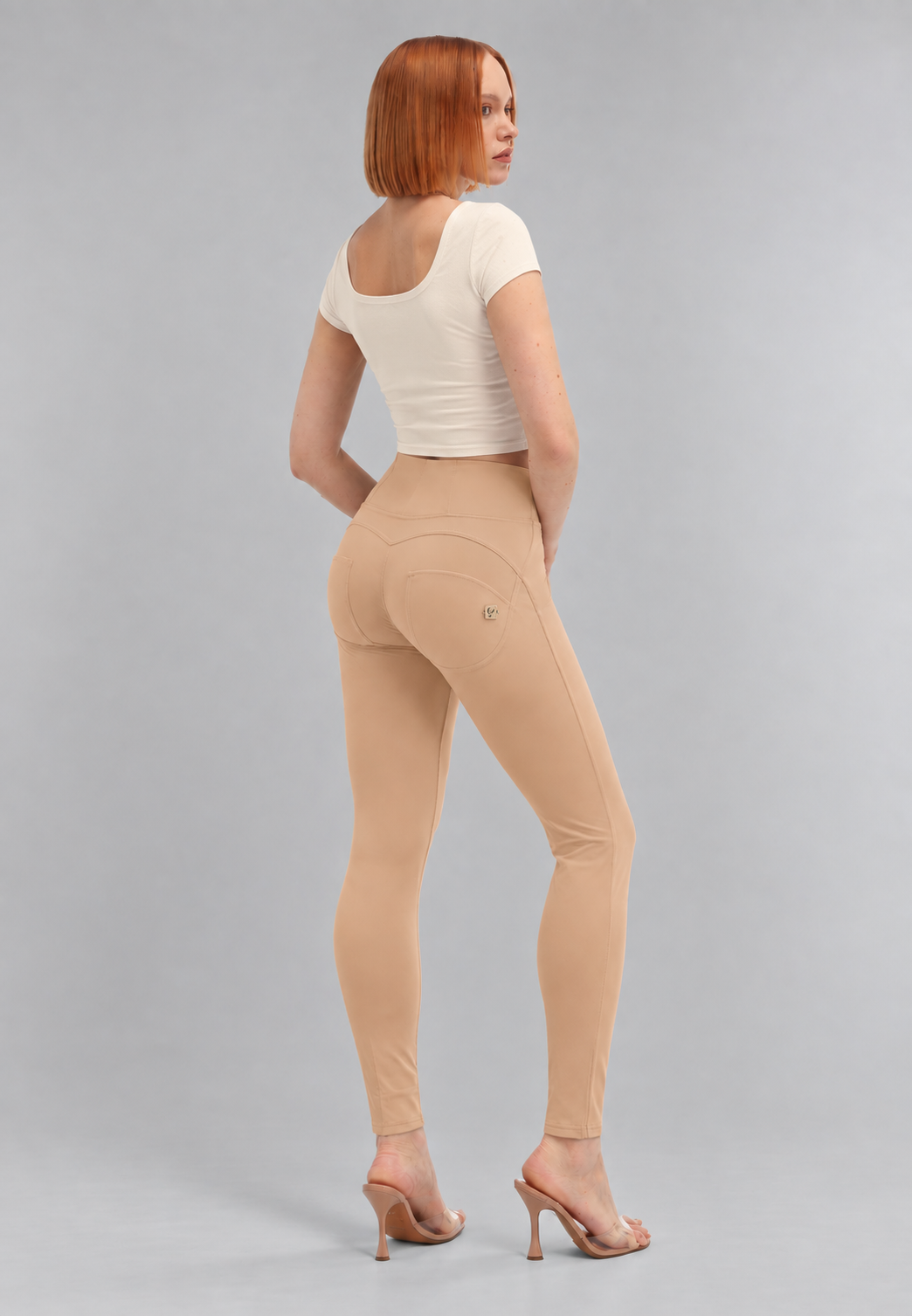 A woman with short red hair is shown from the back wearing beige high-waisted seamless leggings, a white short-sleeve crop top, and nude high-heeled sandals against a light gray studio background.
