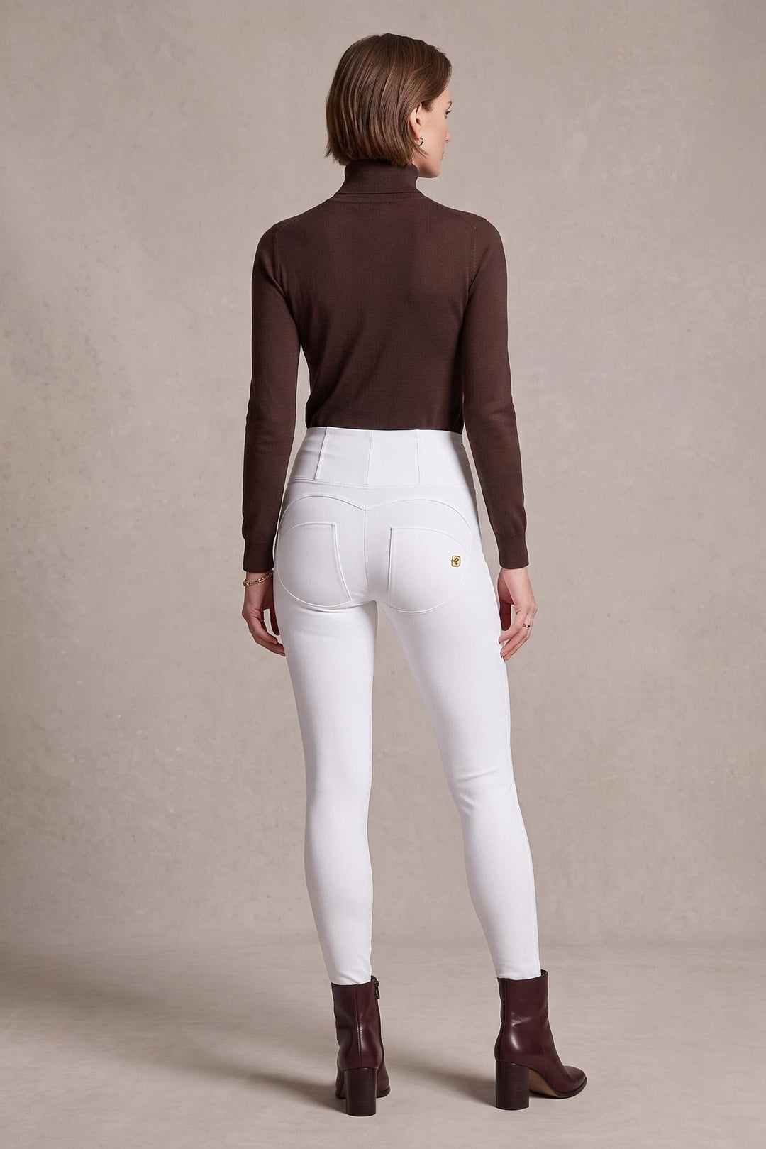 A woman is shown from behind wearing white fitted jeans, a dark brown long-sleeve turtleneck top, and dark brown ankle boots, standing against a neutral textured beige studio backdrop.