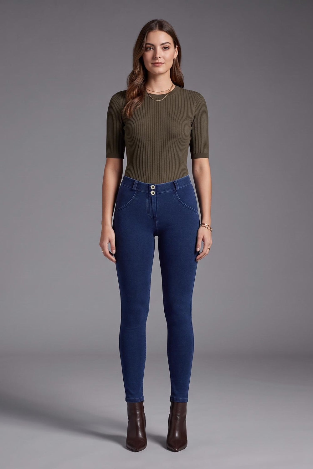 A woman stands against a gray studio background wearing a fitted olive green short-sleeve top, dark blue skinny jeans, and dark brown ankle boots.
