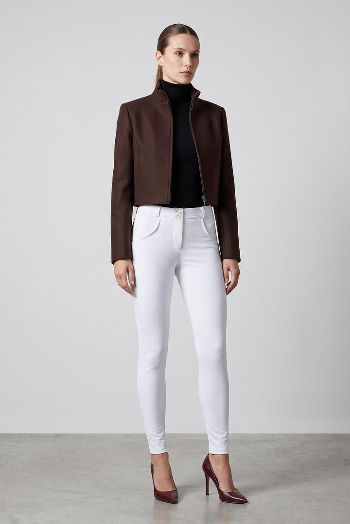 A woman stands in a studio wearing a dark brown cropped jacket over a black turtleneck, white skinny jeans, and burgundy pointed-toe heels against a light gray background.