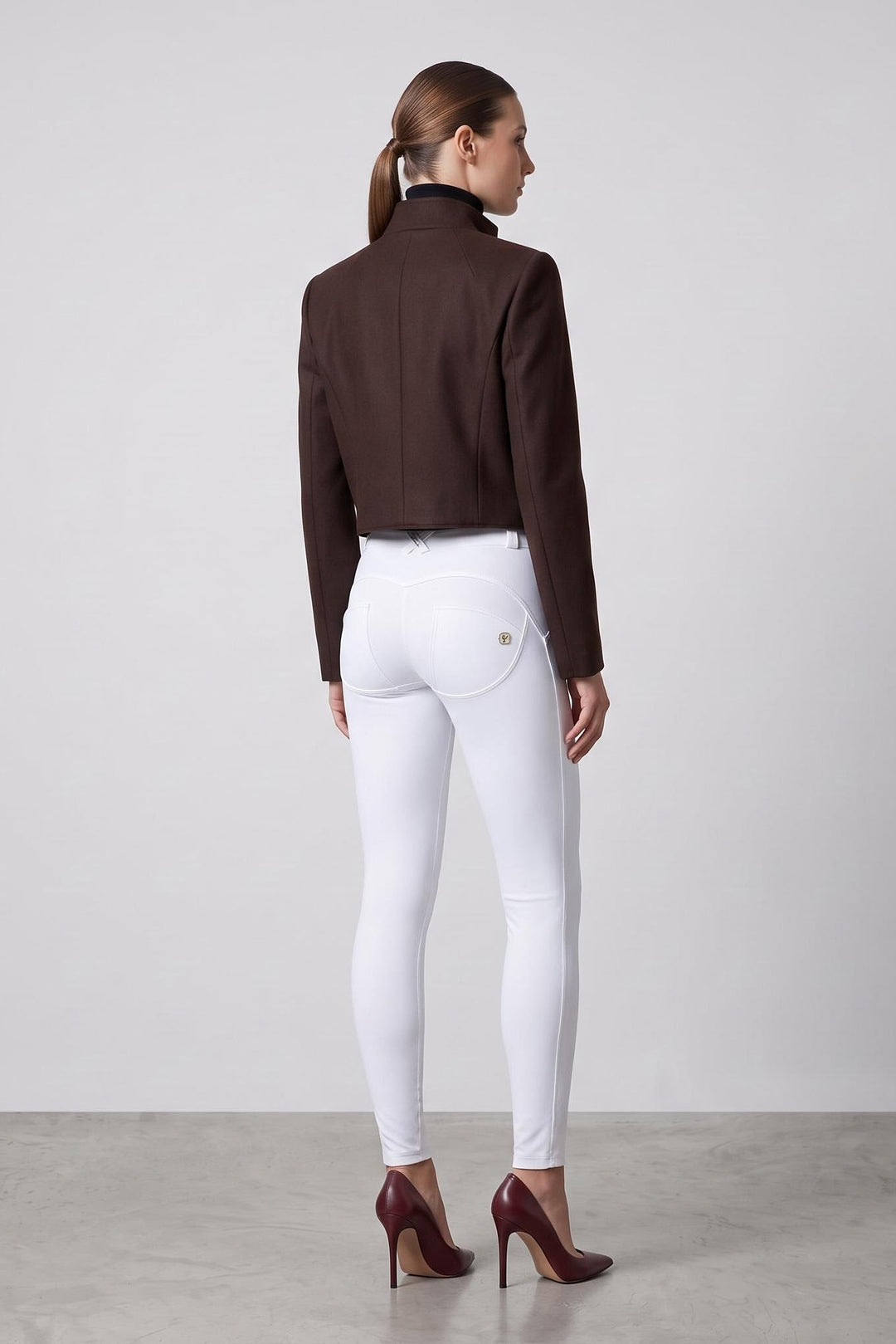 A woman stands with her back to the camera wearing a dark brown cropped jacket, white fitted pants, and burgundy high heels against a light gray studio background.